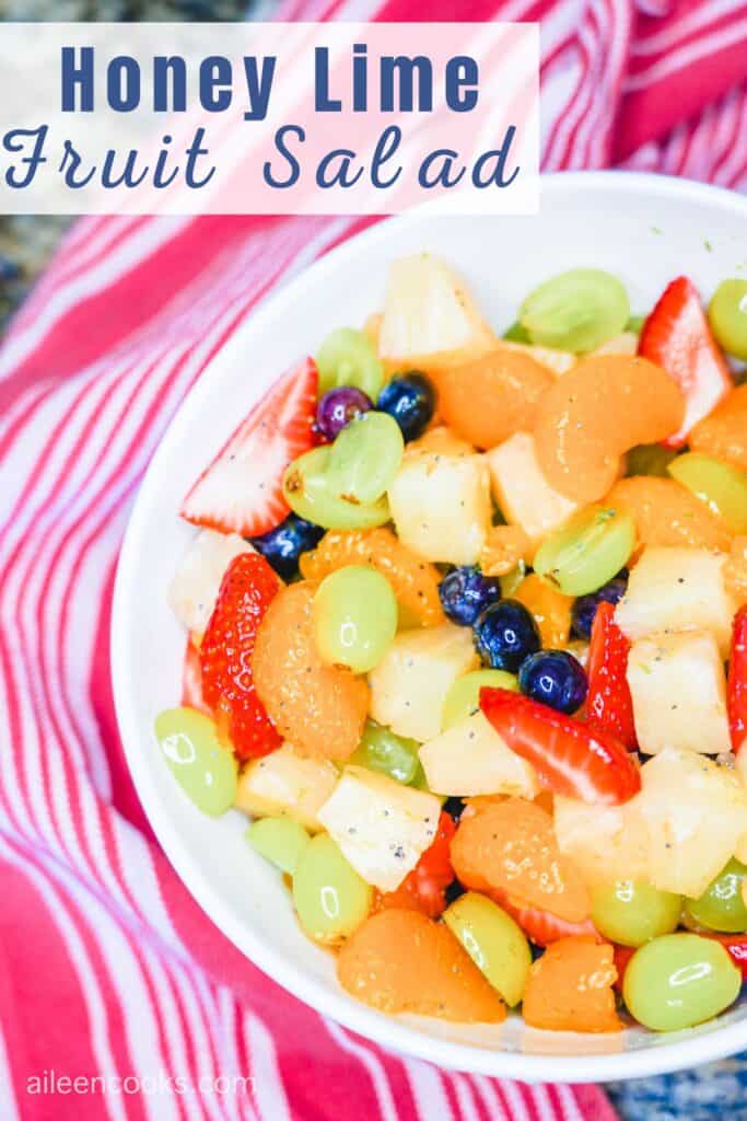 A large white bowl filled with fruit salad with the words "honey lime fruit salad" on the top left hand side of the photo.