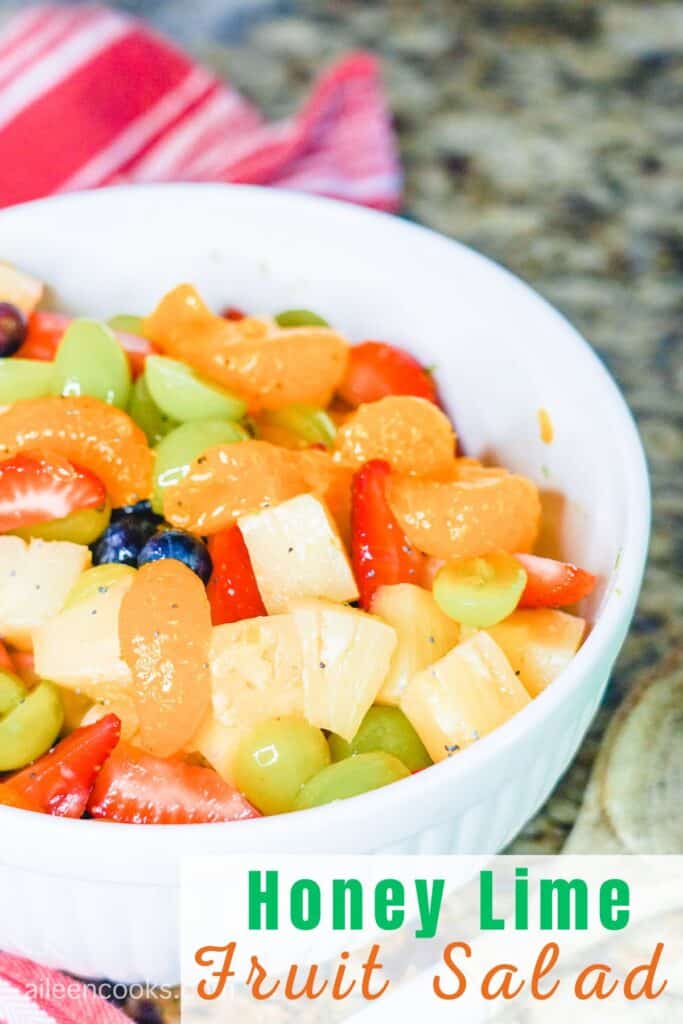 A large white bowl filled with fruit salad with the words "honey lime fruit salad" on the bottom right hand side of the photo.