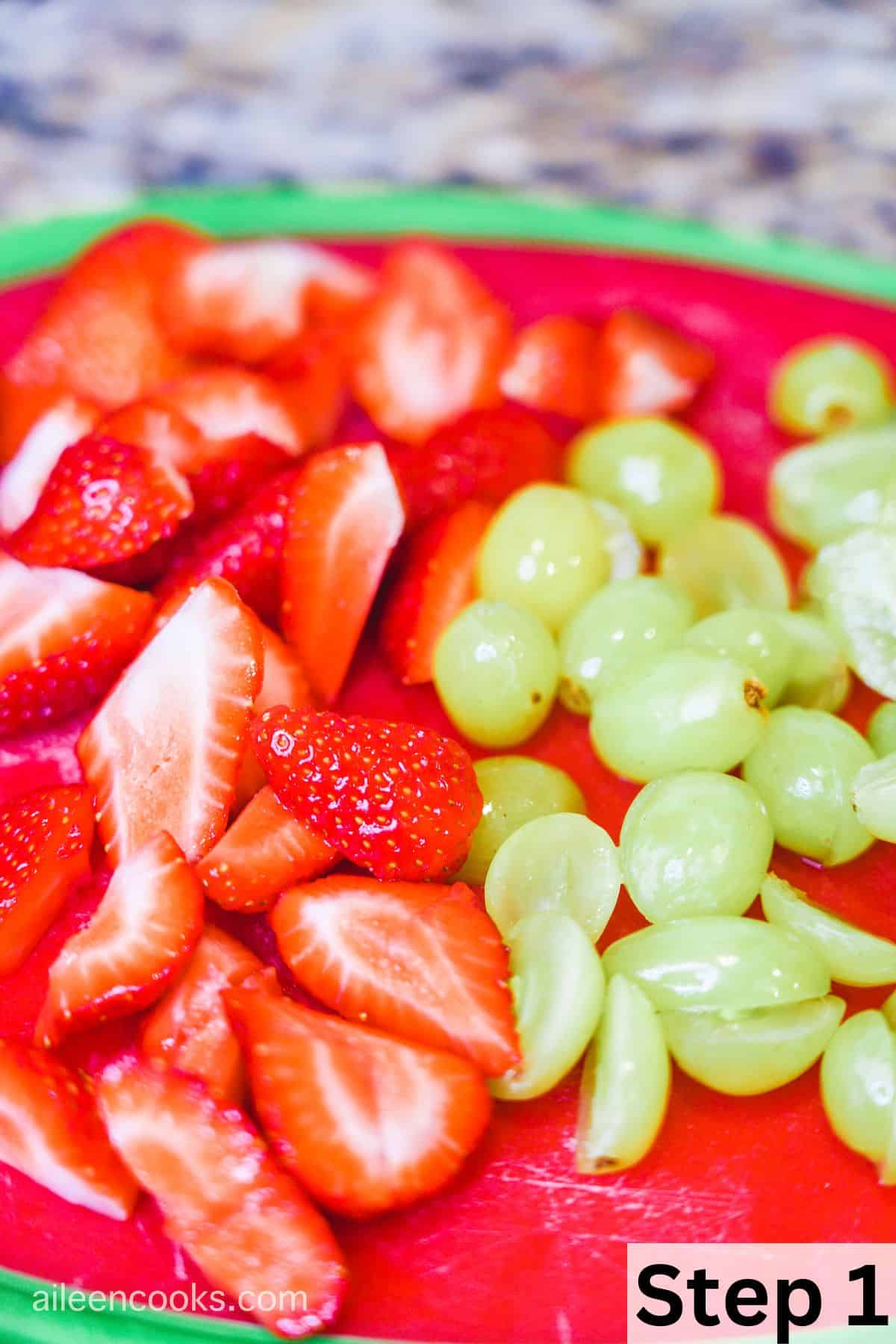 A red cutting board filled with sliced strawberries and green grapes.