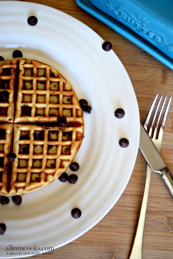 A white plate with a chocolate chip waffle on top and chocolate chips scattered the plate.