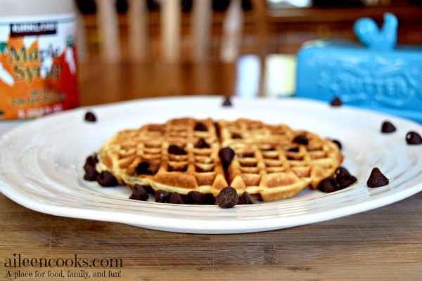 Chocolate chip waffles recipe on a white plate in front of a blue butter dish and maple syrup bottle.