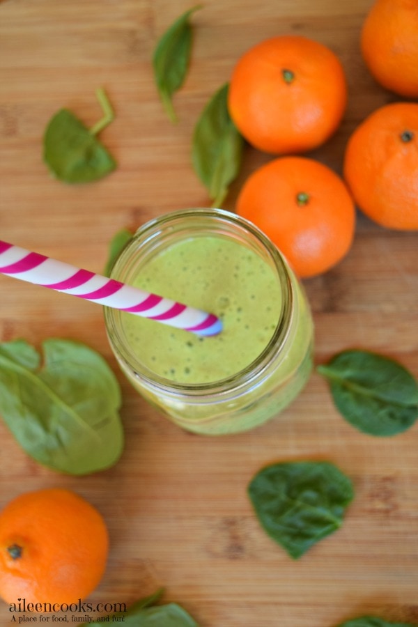 Green smoothie in mason jar with striped straw surrounded by mandarin oranges and baby spinach.