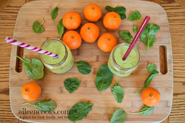 Two orange spinach smoothies in mason jars on a cutting board next to mandarin oranges and baby spinach.