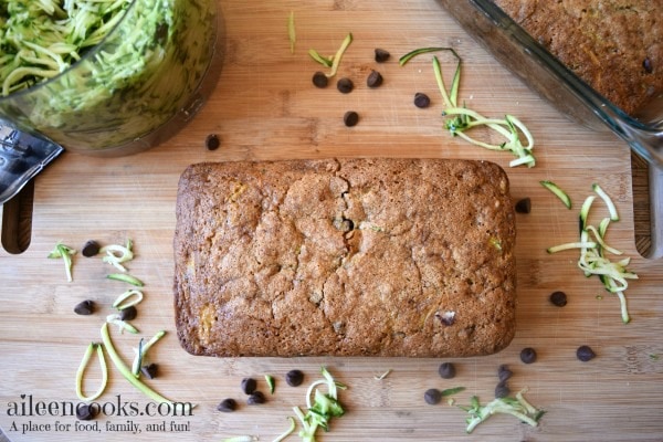A loaf of whole wheat zucchini bread next to a bowl of shredded zucchini and chocolate chips scattered on the surface. 