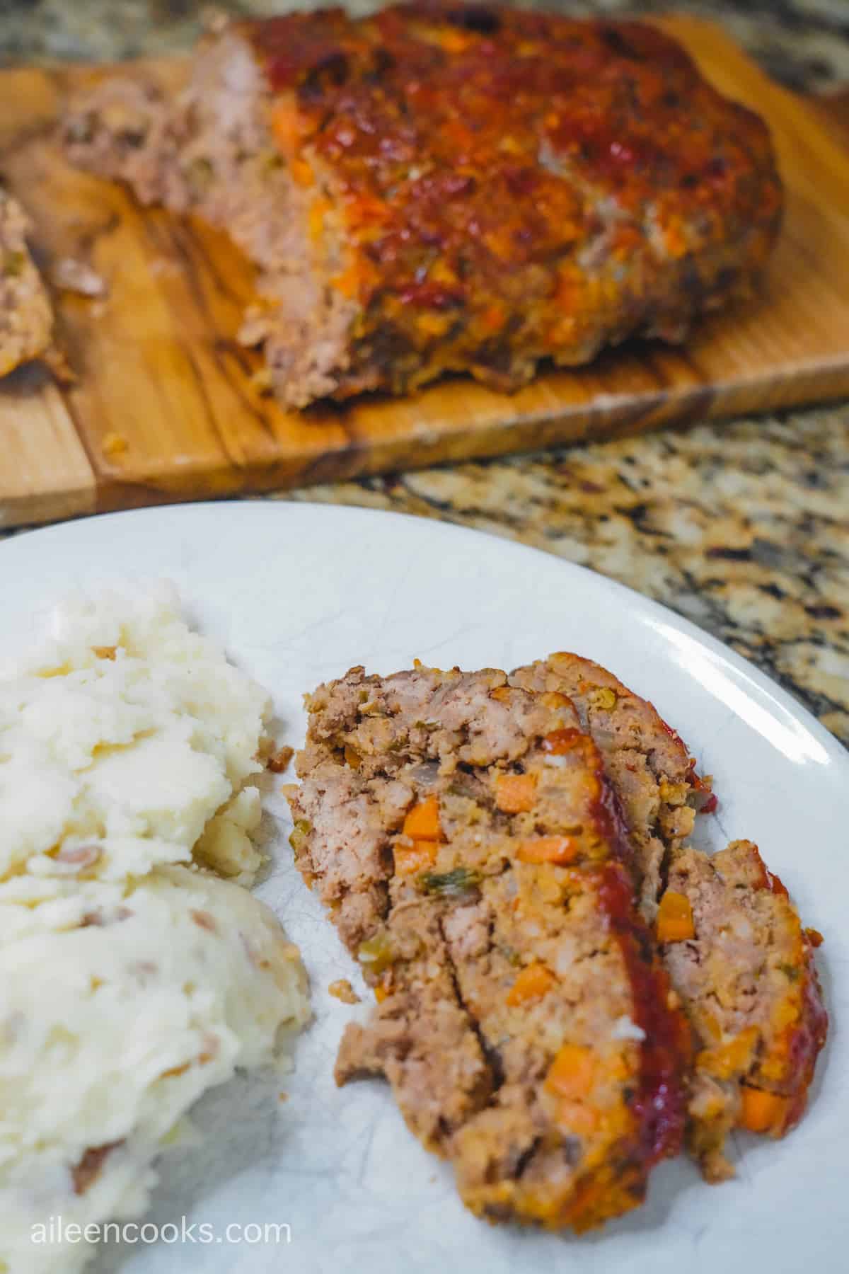 A plate of meatloaf and mashed potatoes in front of a whole meatloaf sliced up on a cutting board.