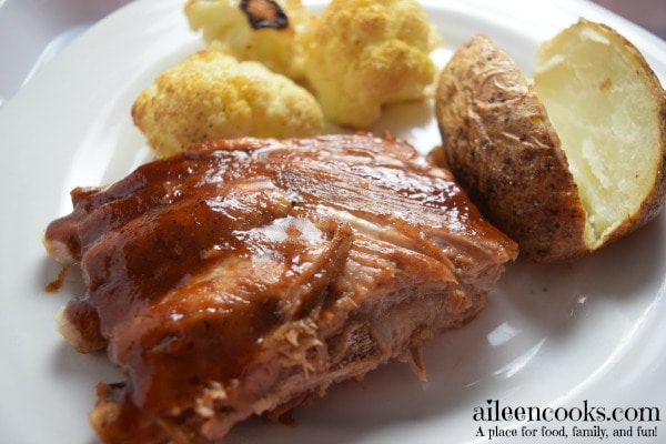 A white plate filled with spare ribs slathered in bbq sauce, baked potato, and roasted cauliflower.