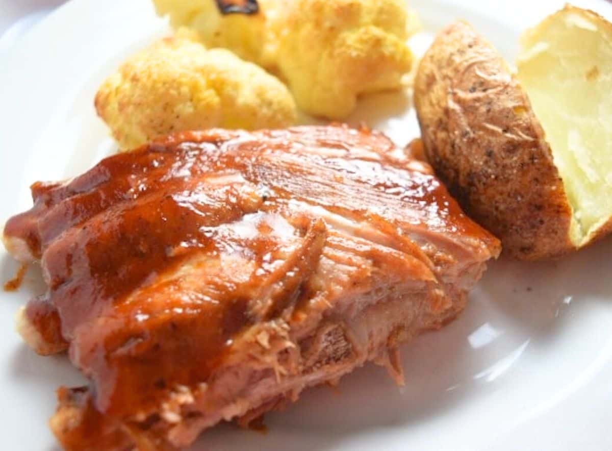 A quarter rack of cooked spare ribs on a white plate next to a baked potato and cauliflower.
