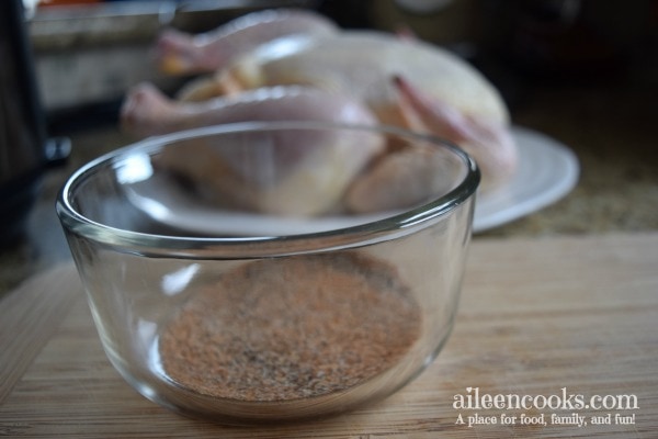 Step-by-step photo showing homemade spice rub in a glass bowl in front of raw whole chicken.