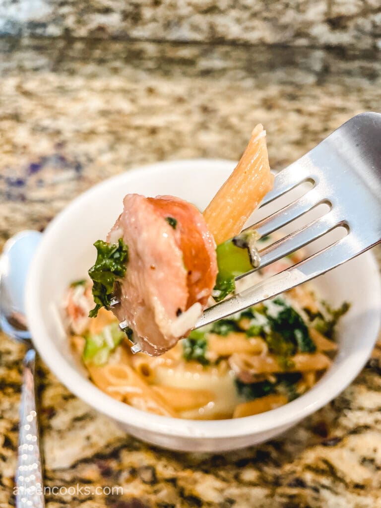 A forkful of pasta, sausage, and kale, above a bowl of pasta.