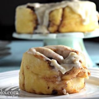Cinnamon roll on a white plate with additional cinnamon rolls on a cake stand in the background.