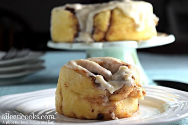 Cinnamon roll on a white plate with additional cinnamon rolls on a cake stand in the background.