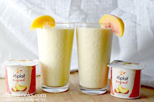 2 glasses of smoothie with slices of peaches on the glass and two containers of Yoplait Harvest Peach yogurt.