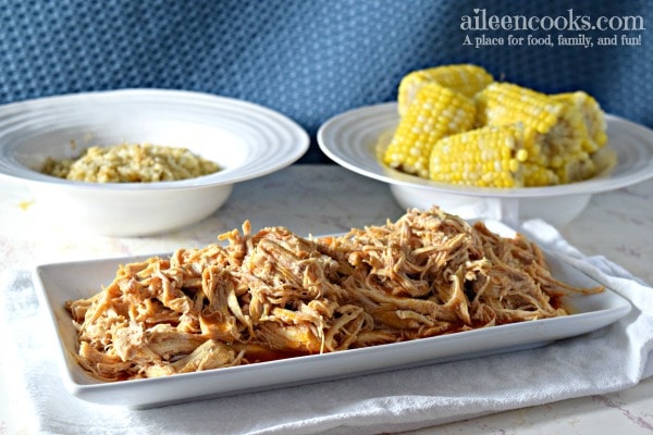 White serving dish with shredded buffalo chicken with bowl of corn in the background.