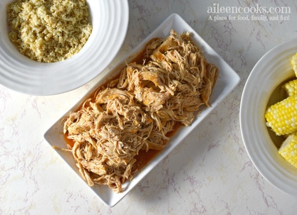 Instant Pot shredded buffalo chicken (made from frozen chicken) on a white platter next to bowl of rice and bowl of corn.