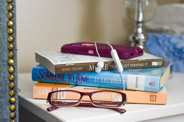 stack of three books, pair of glasses, cell phone, and bluetooth ear buds all sitting on a night stand.