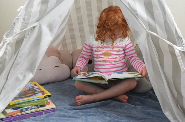 Inside a reading nook filled with soft pillows, books, and a little girl reading to herself.
