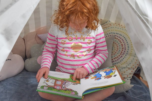 Little girl pointing to a picture in her book, sitting inside a reading nook.