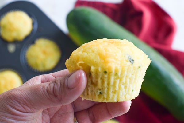 hand holding a cheesy cornbread zucchini muffin with batch of muffins and zucchini in background