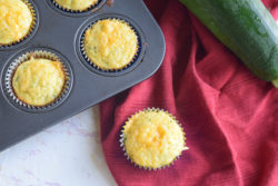 cheesy cornbread zucchini muffin on red cloth napkin next to muffin tin full of muffins and large zucchini