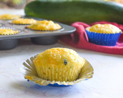 cheesy cornbread zucchini muffin sitting on white counter with muffin tin and zucchini in background