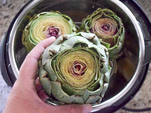 Three raw artichokes in an instant pot. Once is in the process of being placed inside.