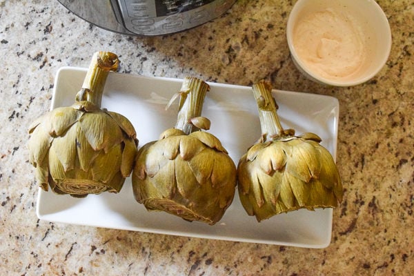 Steamed artichokes on a platter next to dipping sauce and an instant pot.