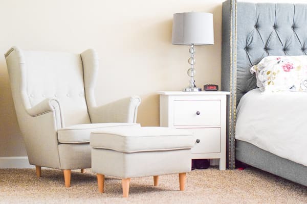 Grey wingback chair, white nightstand with grey lamp and grey bed bed with white floral bedding.