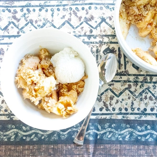 White bowl filled with pressure cooker apple crisp next to a push pan filled with apple crisp.