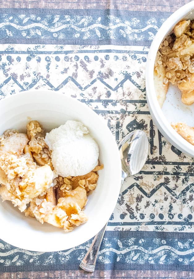 A bowl full of instant pot apple crisp and vanilla ice cream on top of a blue and brown patterned table cloth. 