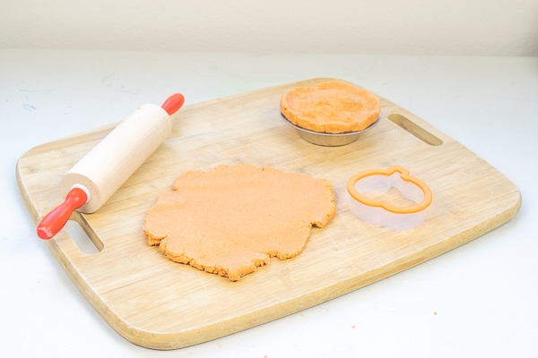 Pumpkin playdough on a cutting board with a rolling pin and pumpkin shaped cookie cutter. 