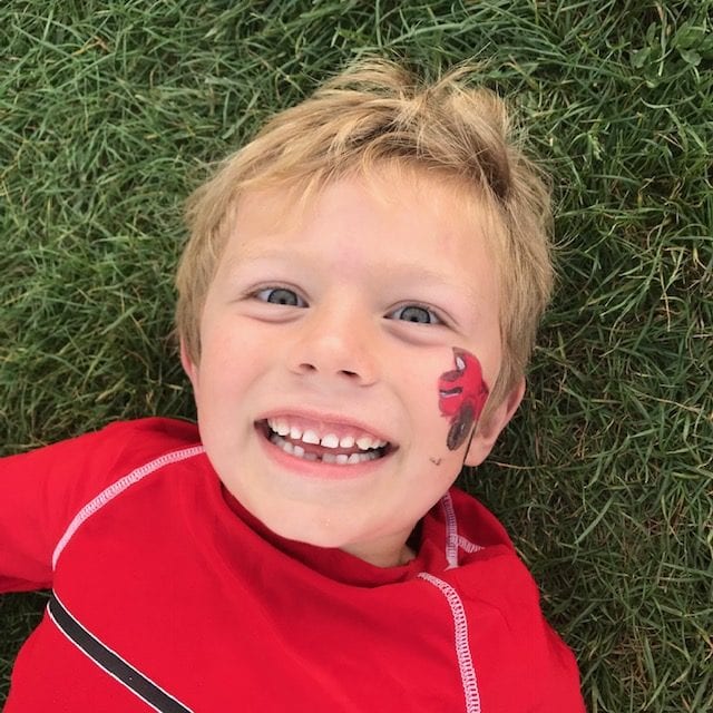 An excited little boy lying in grass with a monster truck painted on his face.