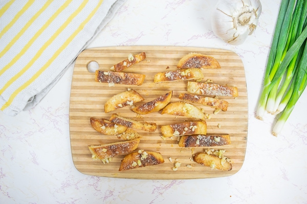 parmesan and garlic crisps on a wooden cutting board next to fresh garlic and scallions.