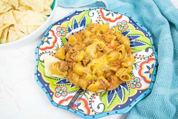 A colorful plate topped with a big portion of instant pot cheesy taco pasta next to a bowl full of tortilla chips.