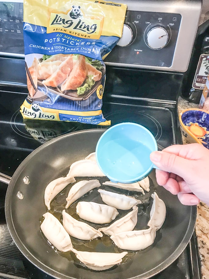 A cup of water being poured into a pan of frozen potstickers. 