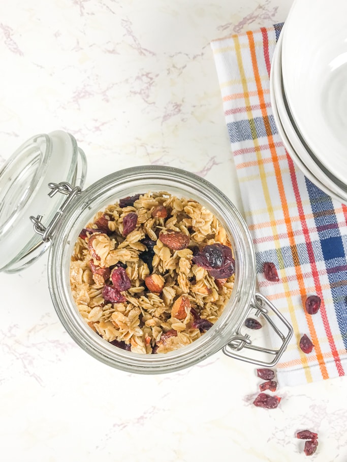 An open jar of instant pot granola next to a pile of dried cranberries and a stack of white bowls.