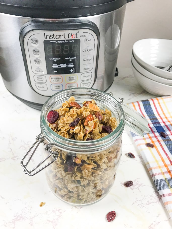 A jar full of homemade granola in front of an instant pot, stack of white bowls, and dish towel.