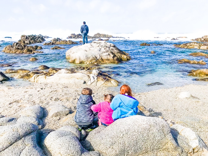 Three children sitting together and watching a man standing on a large rock in the ocean.