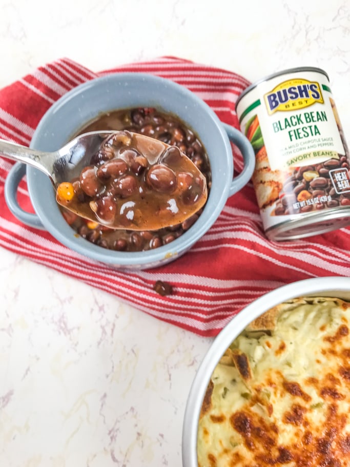 A spoonful of BUSH'S Savory Beans over a bowl of beans and pan of instant pot enchiladas. 