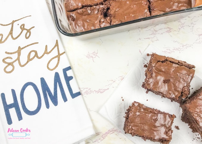 A dish towel that says "Let's stay home" next to a batch of frosted chocolate chunk brownies.