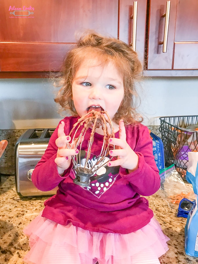 A little girl licking a whisk coated in chocolate frosting.