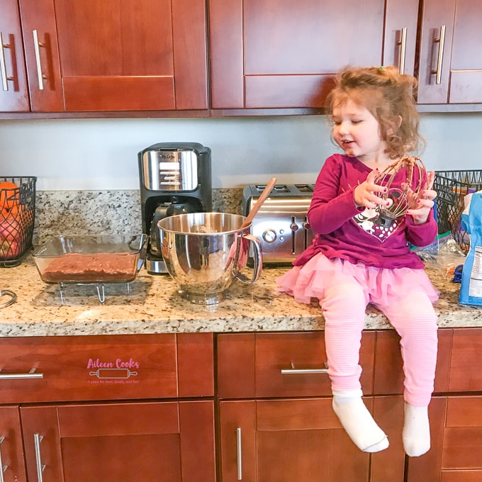 A little girl sitting on a kitchen counter holding a whisk covered in chocolate frosting, next to a batch of chocolate chunk brownies.