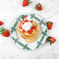 A stack of pancakes topped with strawberries and whipped cream, surrounded by more strawberries.