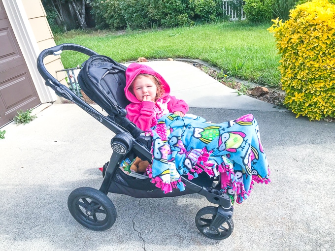 A little girl in a black stroller covered in a pink and blue fleece tie blanket.