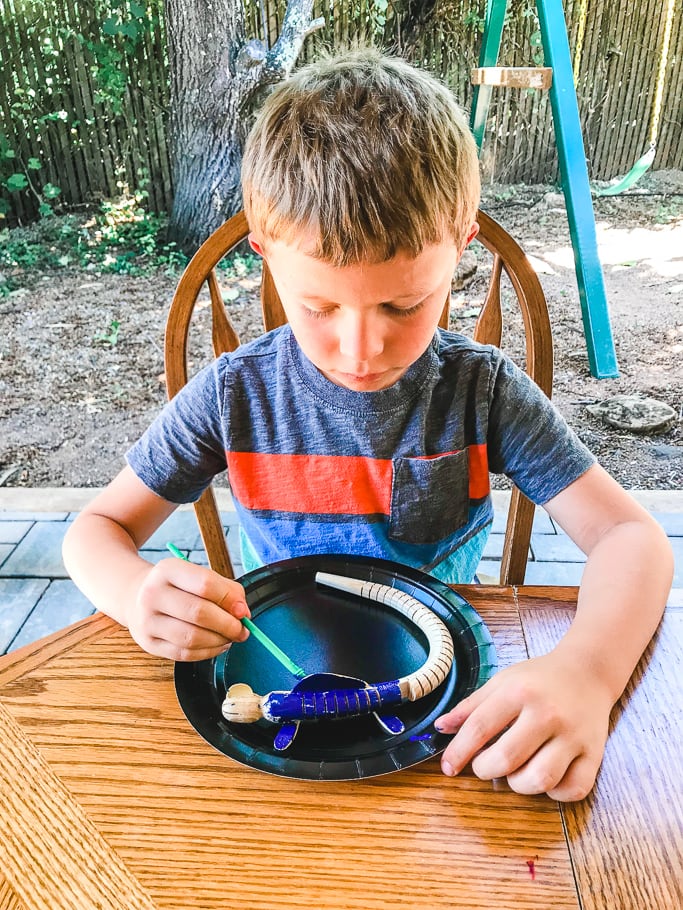 A boy sitting outside painting a wooden lizard.