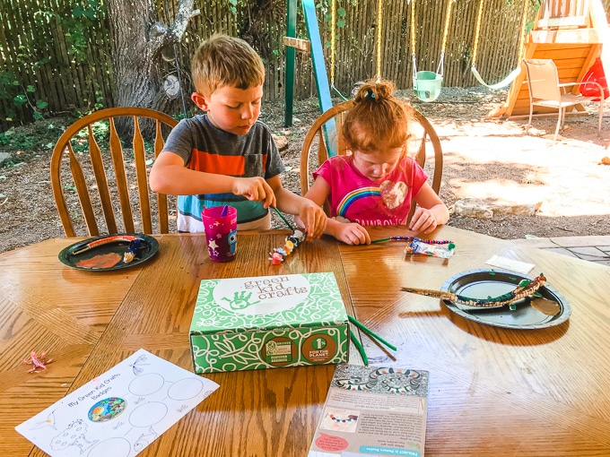 Two kids sitting outside at a wooden table completing projects from their green kid craft subscription box.