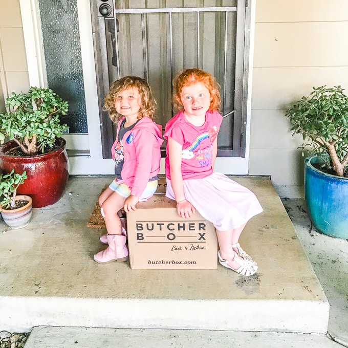 Two girls sitting on a Butcher Box delivery, on their front porch.