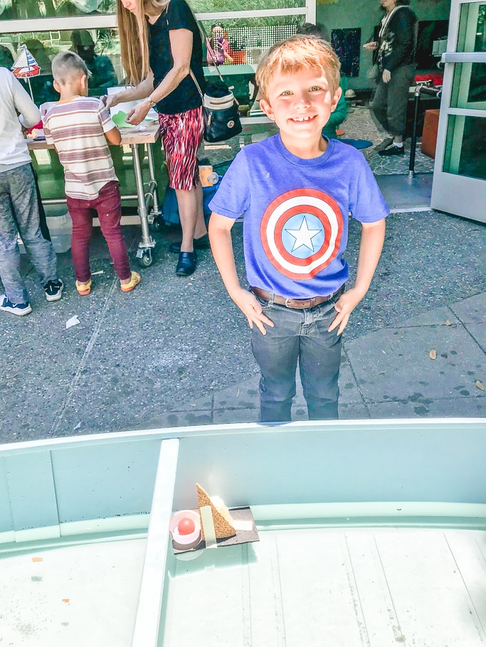 A boy standing next to the boat he built that is floating in water.