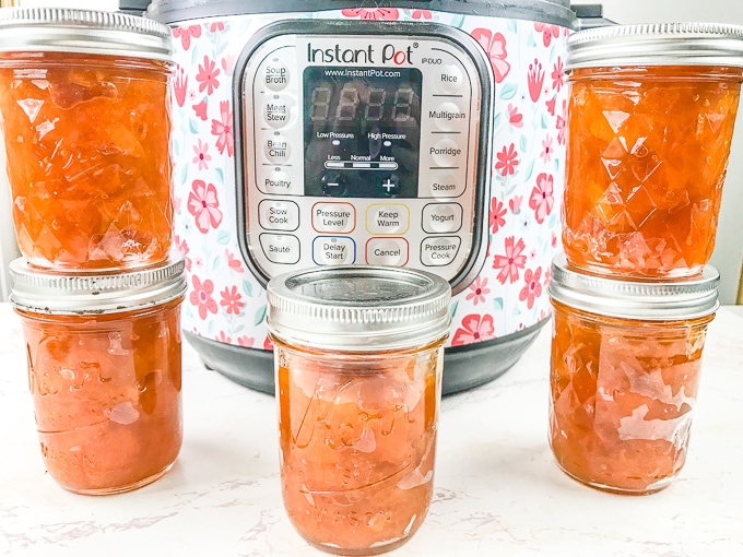 5 jars of instant pot apricot jam stacked up in front of an instant pot with a floral cover.
