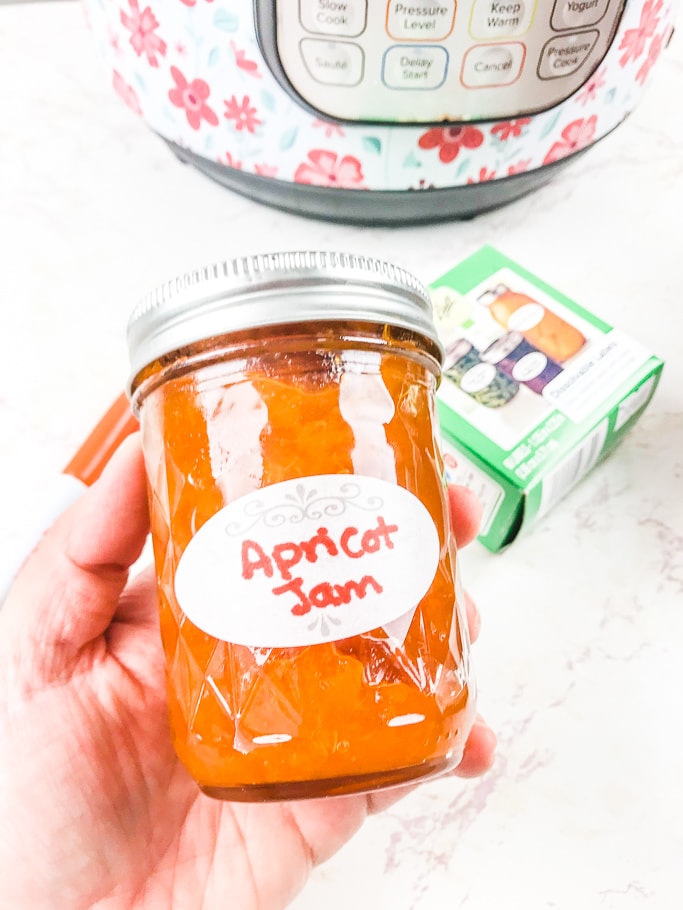 A hand holding a jar of instant pot apricot jam with a white label.