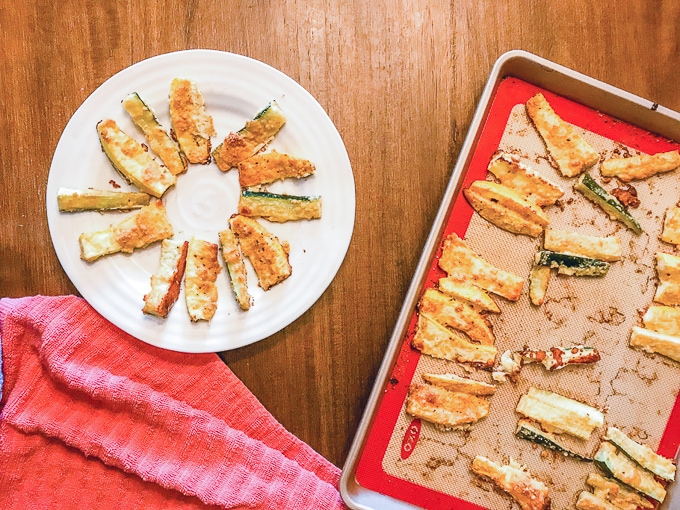 Parmesan zucchini fries arranged in a circle on a white plate next to a cookie sheet full of zucchini fries.